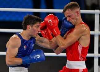 Two men boxing in a white boxing ring. One is wearing red gloves and the other blue. Both gloves have the Paris Olympics logo on them. The boxer in blue is Marco Verde of Mexico and the one in red is Lewis Richardson of the U.K.