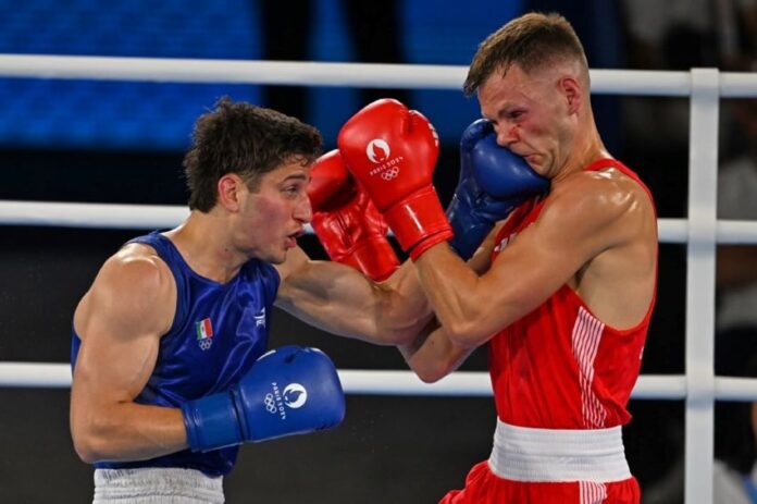 Two men boxing in a white boxing ring. One is wearing red gloves and the other blue. Both gloves have the Paris Olympics logo on them. The boxer in blue is Marco Verde of Mexico and the one in red is Lewis Richardson of the U.K.