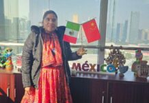 Lorena Ramirez posing while standing in a traditional Raramuri dress and holding small flags from Mexico and China.