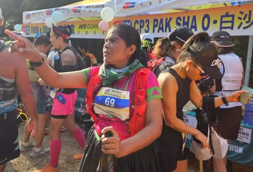 Mexican Raramuri ultramarathoner Lorena Ramirez preparing to race in the Hong Kong Ultramarathon in 2025. She is pointing at something off camera at the starting line while holding a walking stick and dressed in a traditional Raramuri dress.