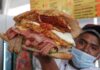 Mexican man holding up an overstuffed torta sandwich for the camera. It's filled with various strips of meat and a fried egg.