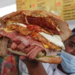 Mexican man holding up an overstuffed torta sandwich for the camera. It's filled with various strips of meat and a fried egg.