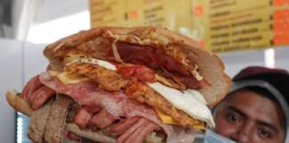 Mexican man holding up an overstuffed torta sandwich for the camera. It's filled with various strips of meat and a fried egg.
