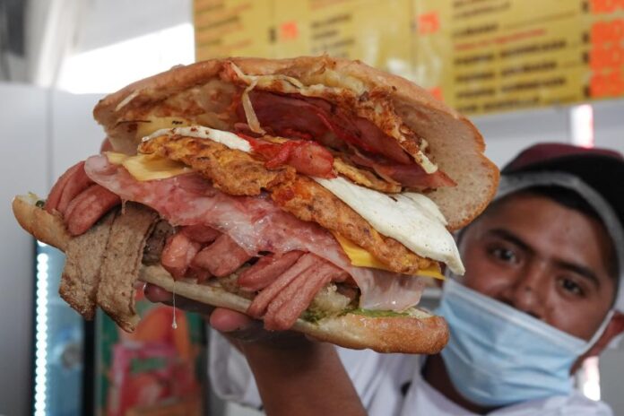 Mexican man holding up an overstuffed torta sandwich for the camera. It's filled with various strips of meat and a fried egg.