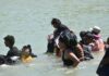 Migrant adults and children walking across the Rio Bravo. The water is up to their waists and shoulders.