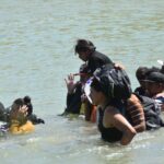 Migrant adults and children walking across the Rio Bravo. The water is up to their waists and shoulders.