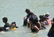 Migrant adults and children walking across the Rio Bravo. The water is up to their waists and shoulders.