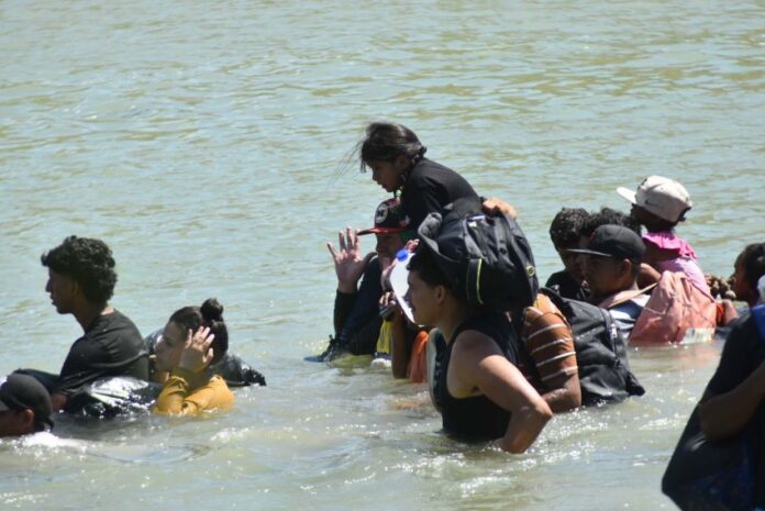 Migrant adults and children walking across the Rio Bravo. The water is up to their waists and shoulders.