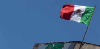 Mexican flag waving in the wind atop a concrete building with Mexico's Federal Electricity Commission logo on the facade in green letters.