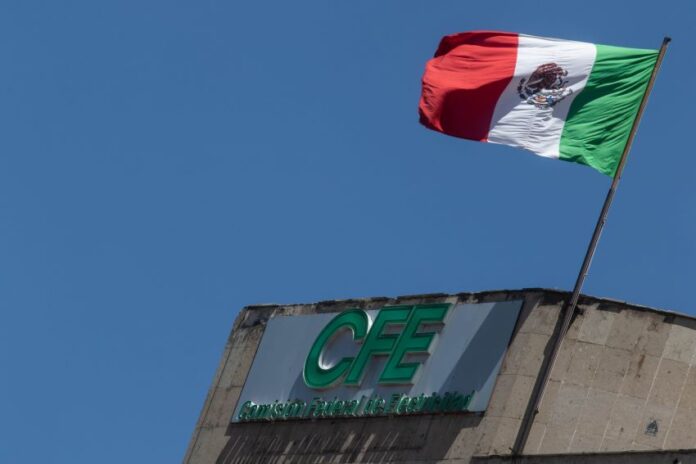 Mexican flag waving in the wind atop a concrete building with Mexico's Federal Electricity Commission logo on the facade in green letters.