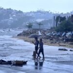 Two people walk under an umbrella on a beach in Acapulco on a rainy day, with storm damaged buildings in the background