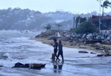 Two people walk under an umbrella on a beach in Acapulco on a rainy day, with storm damaged buildings in the background