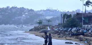 Two people walk under an umbrella on a beach in Acapulco on a rainy day, with storm damaged buildings in the background