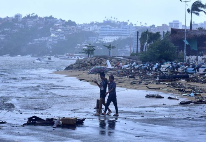 Two people walk under an umbrella on a beach in Acapulco on a rainy day, with storm damaged buildings in the background