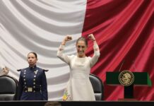 Mexico's President Claudia Sheinbaum in a white dress with embroidered flowers raising her fists in the air in a victorious gesture and smiling on stage in Mexico's Congress. Nearby and to the side, a young female soldier in military dress blues stands watch.