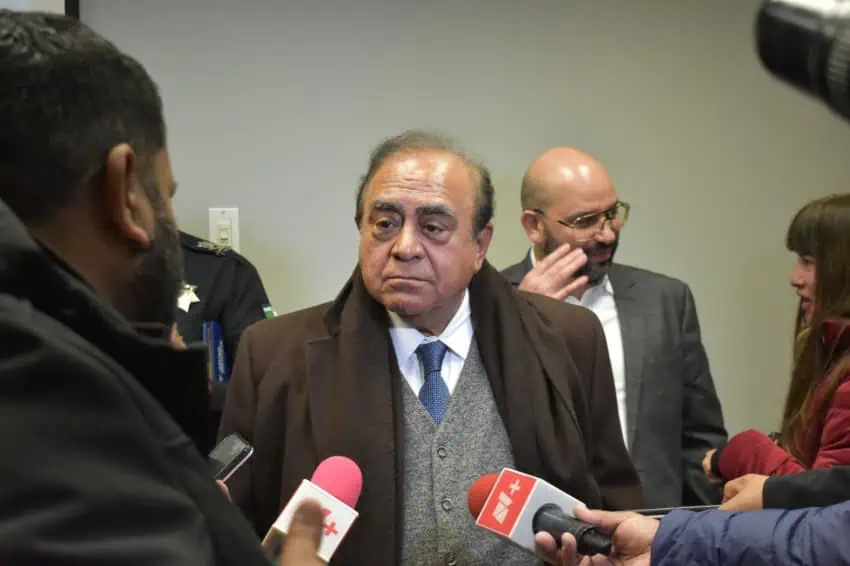 A man standing at a press conference with reporters pointing television microphones at him as he answers questions.