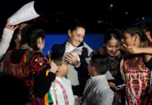 Mexican President Claudia Sheinbaum surrounded by Indigenous young women and children, who are smiling, looking at photos on their phone and clamoring for autographs