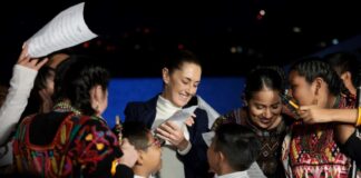 Mexican President Claudia Sheinbaum surrounded by Indigenous young women and children, who are smiling, looking at photos on their phone and clamoring for autographs