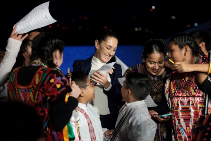 Mexican President Claudia Sheinbaum surrounded by Indigenous young women and children, who are smiling, looking at photos on their phone and clamoring for autographs