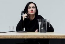 Architect Frida Escobedo sitting at a butcher block table with a tabletop microphone stand, a filled glass pitcher of water. She is sitting with her right hand crooked against the right side of her face and staring off into the distance.