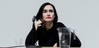 Architect Frida Escobedo sitting at a butcher block table with a tabletop microphone stand, a filled glass pitcher of water. She is sitting with her right hand crooked against the right side of her face and staring off into the distance.