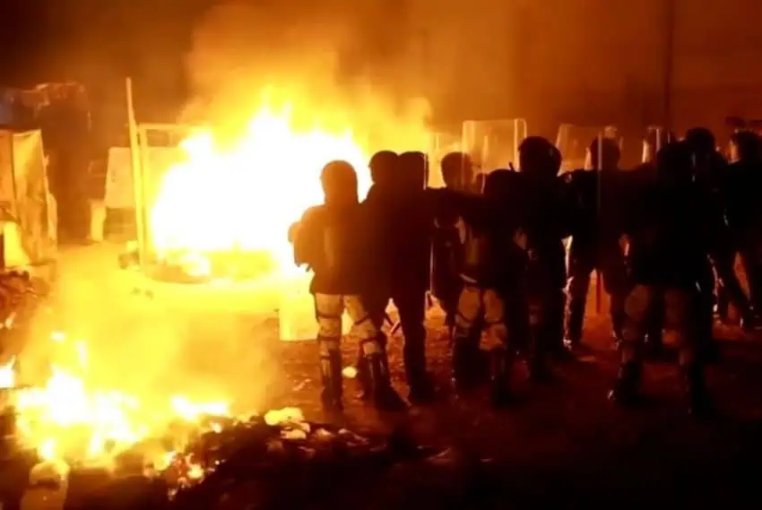 Police with riot shields stand in front of a massive fire