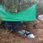 A green tarp held up by stakes in the ground in a grassy, sandy area. Underneath and around the lean-to are clothes and other gear not clear enough in the photo