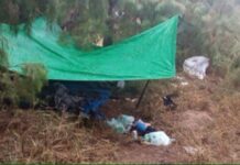 A green tarp held up by stakes in the ground in a grassy, sandy area. Underneath and around the lean-to are clothes and other gear not clear enough in the photo