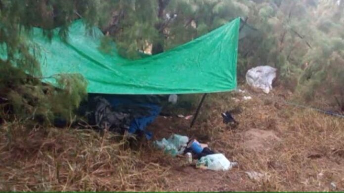 A green tarp held up by stakes in the ground in a grassy, sandy area. Underneath and around the lean-to are clothes and other gear not clear enough in the photo