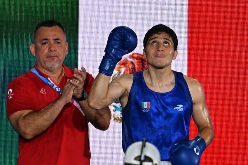 Boxer Marco Verde looking upward to the ceiling in his 2024 Olympic boxing uniform and his boxing gloves on, posing in front of the Mexican flag with an older man in a 2024 Olympics shirt