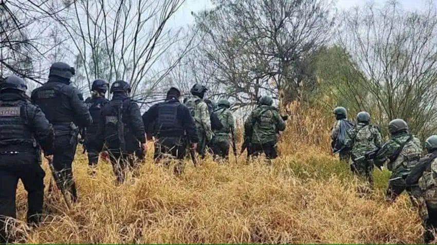 A semicircle of uniformed Mexican soldiers and state police officers from Tamaulipas, Mexico advancing through grassy scrub with tall bare trees in the background.