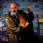 Man in leopard print bathrobe standing in front of wall papered with Mexican newspapers makes devil-horn rock sign with one hand while holding album with another