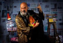 Man in leopard print bathrobe standing in front of wall papered with Mexican newspapers makes devil-horn rock sign with one hand while holding album with another