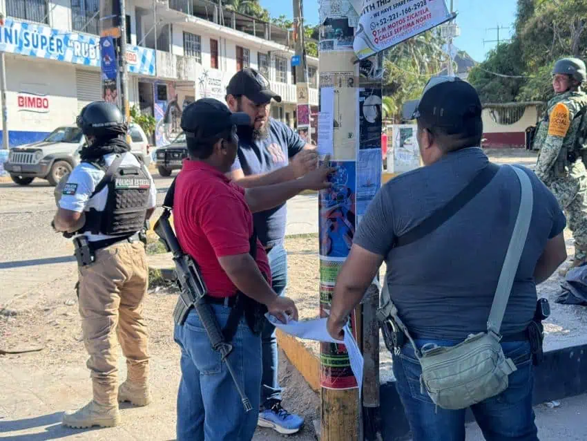 Civilians putting up missing persons poster in a Oaxaca town while a Oaxaca state police officer and a Mexican solder stand guard