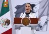 Mexico's President Claudia Sheinbaum standing at the presidential podium during her press conference. Her mouth is open in speech and her eyes are wide and round and she's gripping the podium.