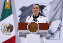 Mexico's President Claudia Sheinbaum standing at the presidential podium during her press conference. Her mouth is open in speech and her eyes are wide and round and she's gripping the podium.