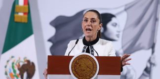 Mexico's President Claudia Sheinbaum standing at the presidential podium during her press conference. Her mouth is open in speech and her eyes are wide and round and she's gripping the podium.