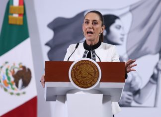 Mexico's President Claudia Sheinbaum standing at the presidential podium during her press conference. Her mouth is open in speech and her eyes are wide and round and she's gripping the podium.