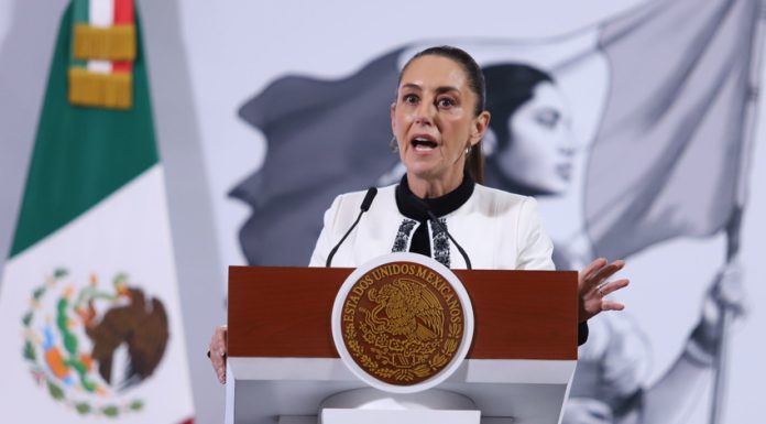 Mexico's President Claudia Sheinbaum standing at the presidential podium during her press conference. Her mouth is open in speech and her eyes are wide and round and she's gripping the podium.