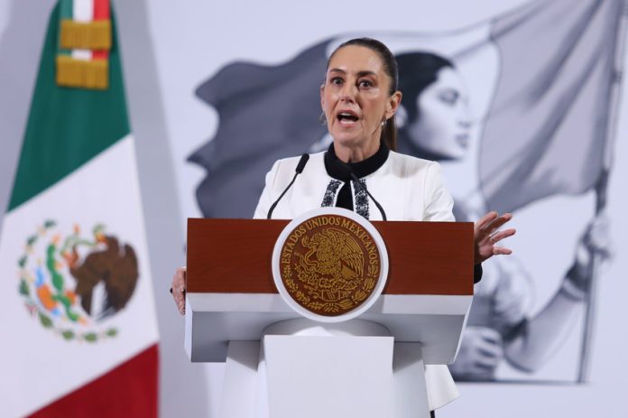 Mexico's President Claudia Sheinbaum standing at the presidential podium during her press conference. Her mouth is open in speech and her eyes are wide and round and she's gripping the podium.