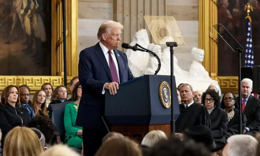 Donald J. Trump giving his inaugural address in January 2025, standing at the presidential podium with distinguished audience members sitting around the podium