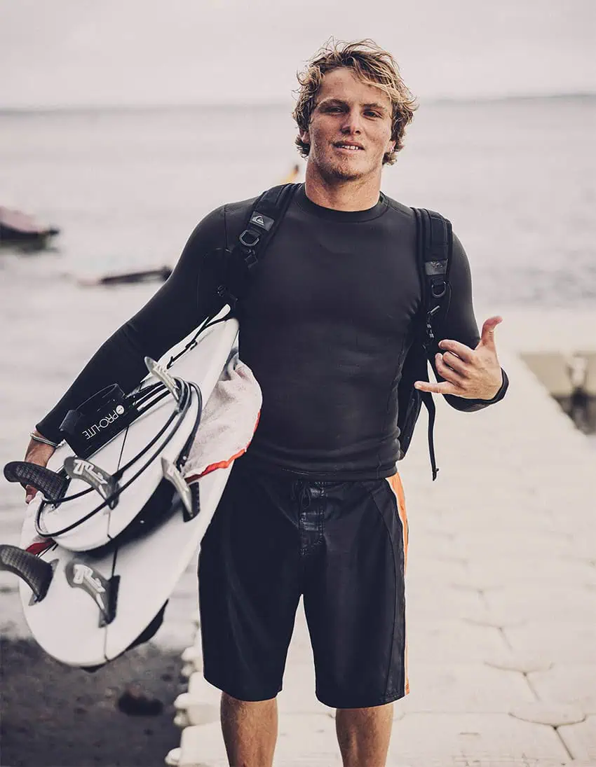 Alan Cleland flashes a hang loose sign at the camera while carrying surfboards by the ocean