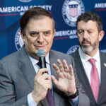 Marcelo Ebrard speaks into a microphone in front of a banner showing the American Society of Mexico logo and name