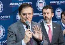 Marcelo Ebrard speaks into a microphone in front of a banner showing the American Society of Mexico logo and name