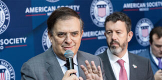 Marcelo Ebrard speaks into a microphone in front of a banner showing the American Society of Mexico logo and name