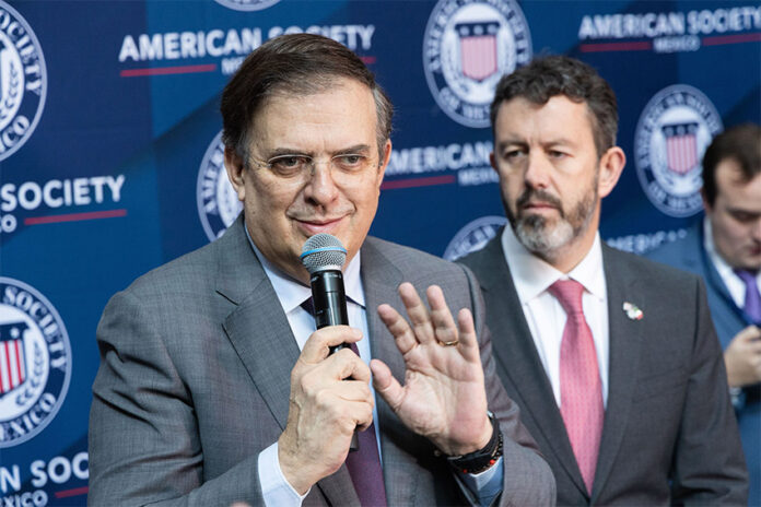 Marcelo Ebrard speaks into a microphone in front of a banner showing the American Society of Mexico logo and name