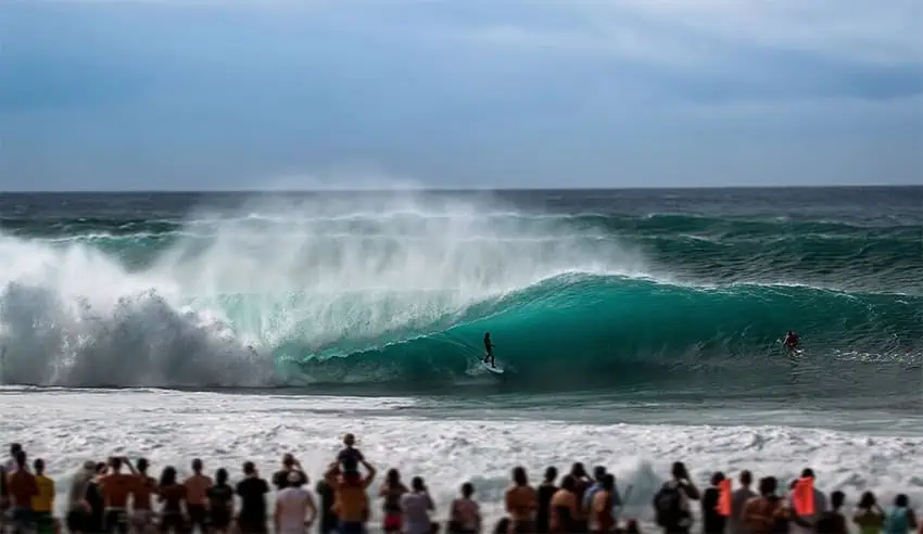 A surfer rides a double-overhead wave at Pipeline while a crowd looks on from the beach