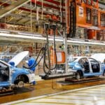 White and blue car frames moving on an assembly line in an expansive factory hangar.