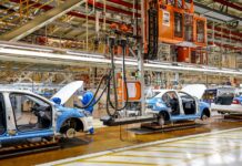 White and blue car frames moving on an assembly line in an expansive factory hangar.