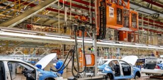 White and blue car frames moving on an assembly line in an expansive factory hangar.